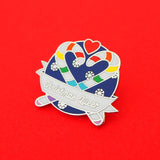 Round enamel pin showing two rainbow striped candy canes crossed at an angle which makes their hooks form a heart shape. There is a small red heart above them and the background is snowflakes on dark blue. There is a ribbon banner at the bottom which reads Christmas Queer. The pin is on a bright red background.