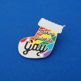 Enamel pin in the shape of a Christmas stocking with rainbow stripes, it reads Making Yuletide Gay in black script. The word Gay is larger and has star sparkles. The pin is on a blue background.