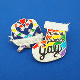 Two enamel pins. The first is round, showing two rainbow striped candy canes crossed at an angle which makes their hooks form a heart shape. There is a small red heart above them and the background is snowflakes on dark blue. There is a ribbon banner at the bottom which reads Christmas Queer. The second is in the shape of a Christmas stocking with rainbow stripes, it reads Making Yuletide Gay. The word Gay is larger and has star sparkles. The pins are on a blue background.