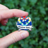 Round enamel pin showing two rainbow striped candy canes crossed at an angle which makes their hooks form a heart shape. There is a small red heart above them and the background is snowflakes on dark blue. There is a ribbon banner at the bottom which reads Christmas Queer. The pin is held between a thumb and forefinger.