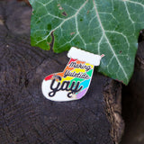 Enamel pin in the shape of a Christmas stocking with rainbow stripes, it reads Making Yuletide Gay in black script. The word Gay is larger and has star sparkles. The pin is resting on a log next to a leaf.