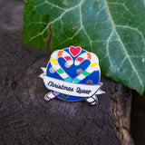 Round enamel pin showing two rainbow striped candy canes crossed at an angle which makes their hooks form a heart shape. There is a small red heart above them and the background is snowflakes on dark blue. There is a ribbon banner at the bottom which reads Christmas Queer. The pin is resting on a log next to a leaf.