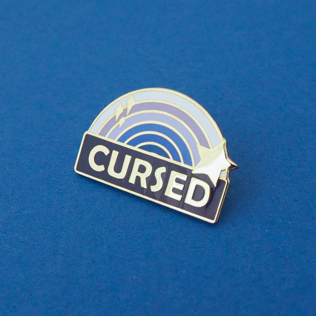 Enamel pin with a blue ombre rainbow above a dark blue banner which has the word Cursed in silver capital letters. The rainbow is decorated with a silver star and sparkles. The pin is on a blue background.