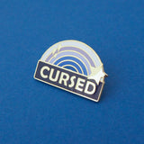 Enamel pin with a blue ombre rainbow above a dark blue banner which has the word Cursed in silver capital letters. The rainbow is decorated with a silver star and sparkles. The pin is on a blue background.