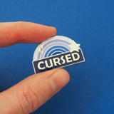 Enamel pin with a blue ombre rainbow above a dark blue banner which has the word Cursed in silver capital letters. The rainbow is decorated with a silver star and sparkles. The pin is being held between a thumb and forefinger.
