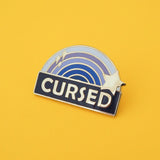 Enamel pin with a blue ombre rainbow above a dark blue banner which has the word Cursed in silver capital letters. The rainbow is decorated with a silver star and sparkles. The pin is on a yellow background.