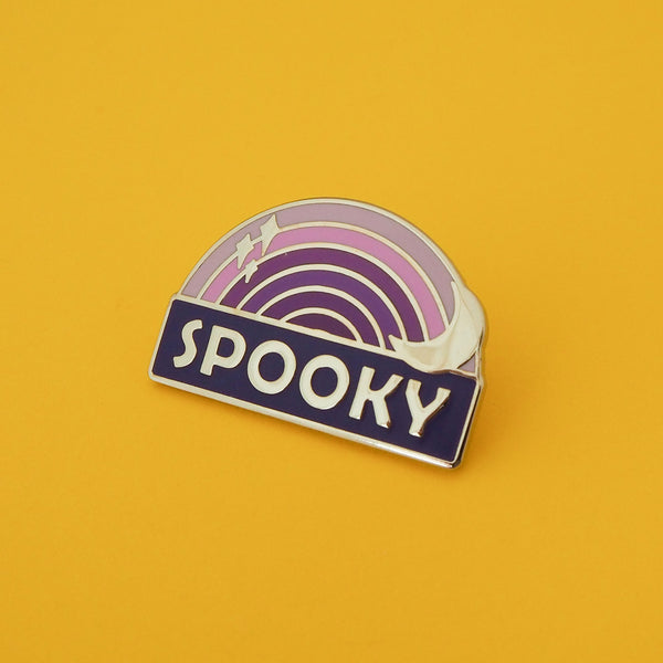 Enamel pin with a purple ombre rainbow above a dark purple banner which reads Spooky in silver capital letters. The rainbow is decorated with a silver crescent moon and sparkles. The pin is shown on a yellow background.