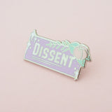 A rectangular enamel pin that reads Dissent in thick silver capital letters on a lavender background. A pastel pink flower with light green leaves and fern is bursting from the top right corner. There are white sparkles in the top left and bottom right corners. The pin is on a pastel pink background.