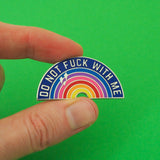 Rainbow enamel pin that reads Do Not Fuck With Me in silver capital letters on a dark blue arch. The colourful rainbow beneath has three silver sparkles in the upper left. The pin is being held between a forefinger and thumb in front of a bright geen background.
