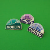 Three enamel pins. One has a green ombre rainbow above a dark green banner with the word Goblin in capital letters. The rainbow shows green mountains with silver caps and silver sparkles. The second is a purple ombre rainbow above a dark purple banner which reads Spooky in capital letters. The rainbow has a silver crescent moon and sparkles. The last is a blue ombre rainbow above a dark blue banner with the word Cursed in capital letters. The rainbow is decorated with a silver star and sparkles.