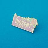 A rectangular enamel pin that reads Dissent in thick silver capital letters on a lavender background. A pastel pink flower with light green leaves and fern is bursting from the top right corner. There are white sparkles in the top left and bottom right corners. The pin is on a blue background.