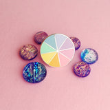 Round enamel pin, split into eight sections. Each section is filled with a bright rainbow colour, replicating a colour wheel. The pin is on a light pink background surrounded by shimmering purple glass pebbles.