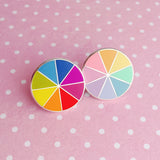Two round enamel pins, split into eight sections. Each section is filled with a bright rainbow colour on the left pin and pastel rainbow colours on the right pin, replicating a colour wheel. The pins are on a light pink, white-dotted background.