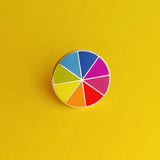 Round enamel pin, split into eight sections.  Each section is filled with a bright rainbow colour, replicating a colour wheel. The pin is shown on a yellow background.