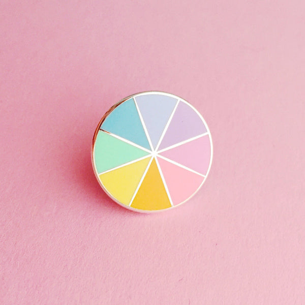 Round enamel pin, split into eight sections. Each section is filled with a pastel rainbow colour, replicating a colour wheel. The pin is on a light pink background.