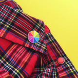 Round enamel pin, split into eight sections. Each section is filled with a bright rainbow colour, replicating a colour wheel. It is pinned to the collar of a red flannel shirt.