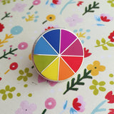 Round enamel pin, split into eight sections. Each section is filled with a bright rainbow colour, replicating a colour wheel. The pin is on a flowery background.