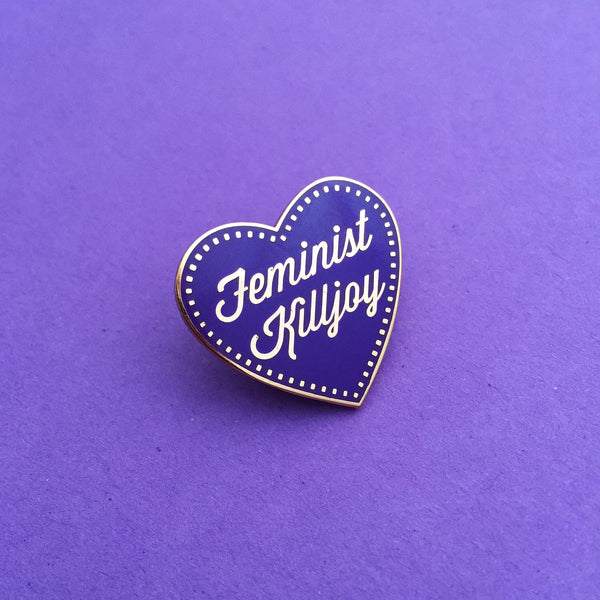 A deep purple enamel pin in the shape of a heart. The words Feminist Killjoy are written in rose gold in a flowing script. Rose gold dots run along the inside border of the heart. The pin is shown on a purple background.
