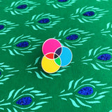 Enamel pin with three overlapping circles. The circles are filled cyan, magenta, and yellow, with the colours mixing in the areas where they overlap. The pin is on a peacock-feather patterned background.